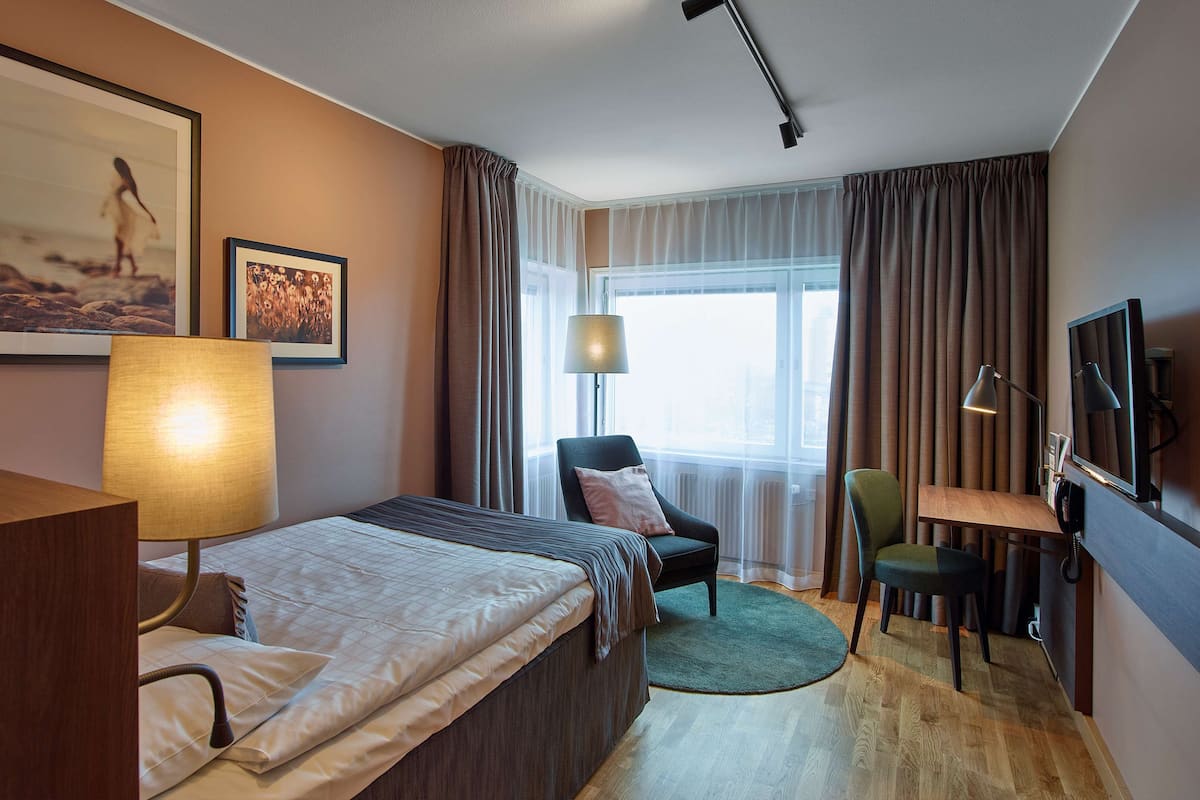 Habitación de hotel con una cama, un escritorio, una silla, una lámpara, un televisor y dos cuadros en la pared.