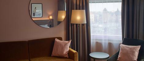A hotel room with a sofa, armchair, round mirror, and a view of the city through a window.