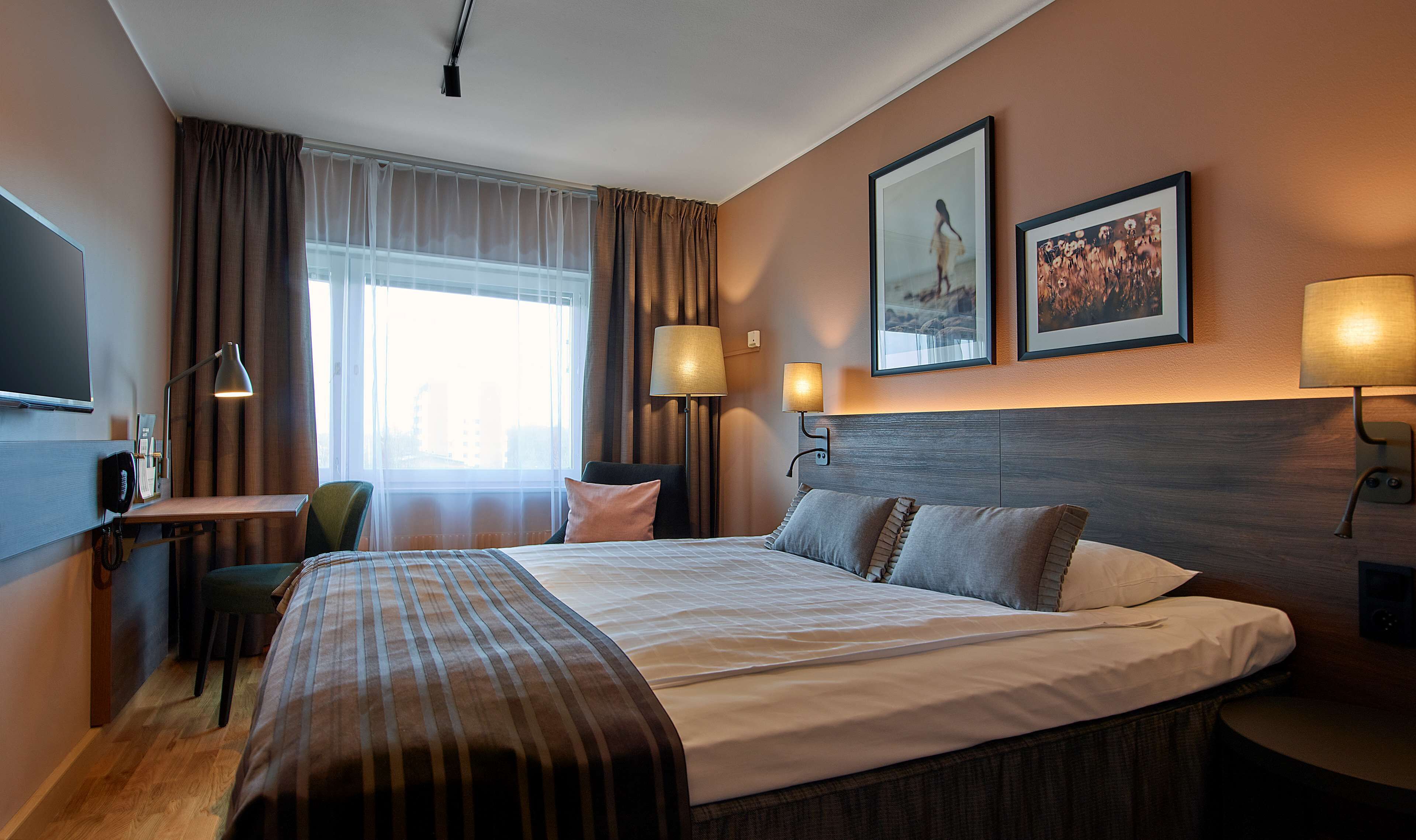 A hotel room with a large bed, two bedside lamps, a desk with a telephone, a chair, and a large window with curtains.