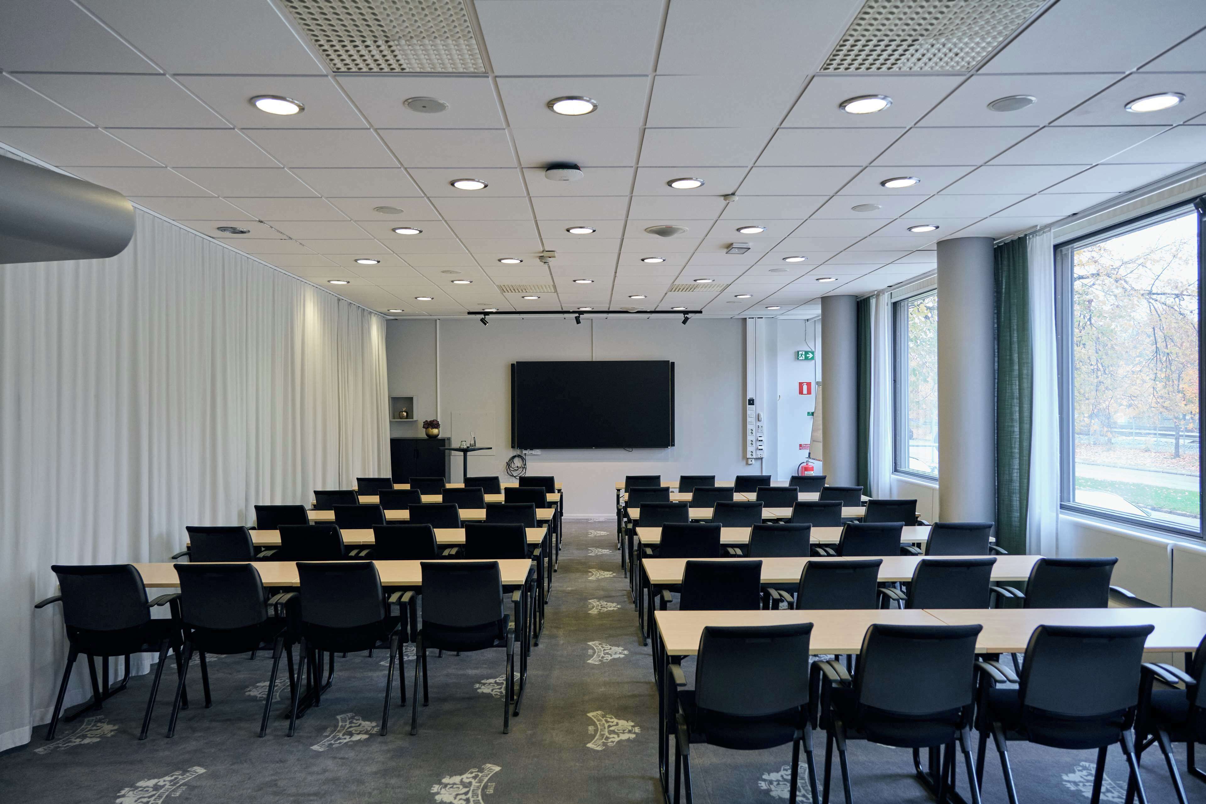 A conference room with rows of tables and chairs, a large screen, and windows with a view of trees.