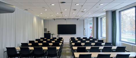 A conference room with rows of tables and chairs, a large screen, and windows with a view of trees.