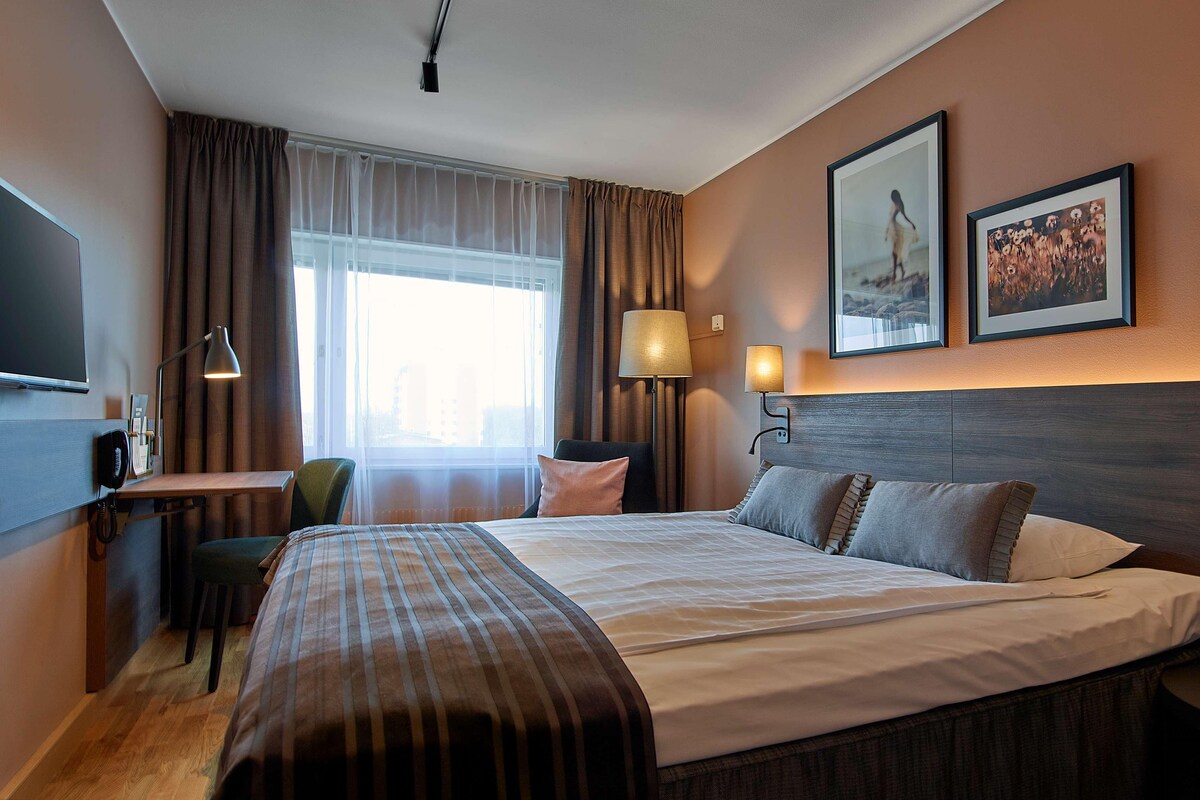 Habitación de hotel con una cama grande, un escritorio con silla, televisión y dos lámparas de pared.