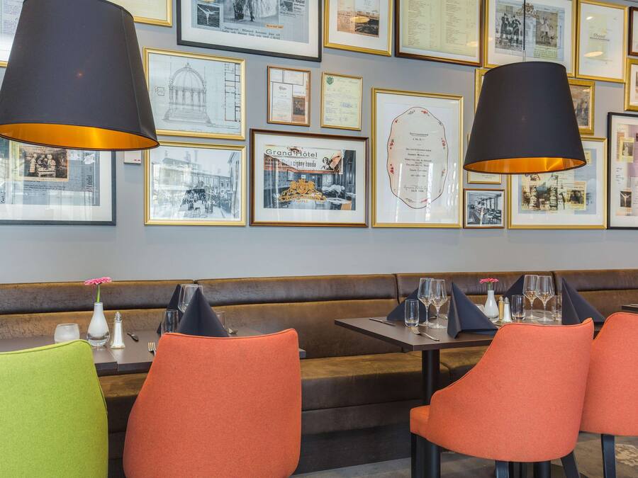 A restaurant interior with a long counter, orange and green upholstered chairs, black and gold framed photographs on the wall, and black pendant lighting.