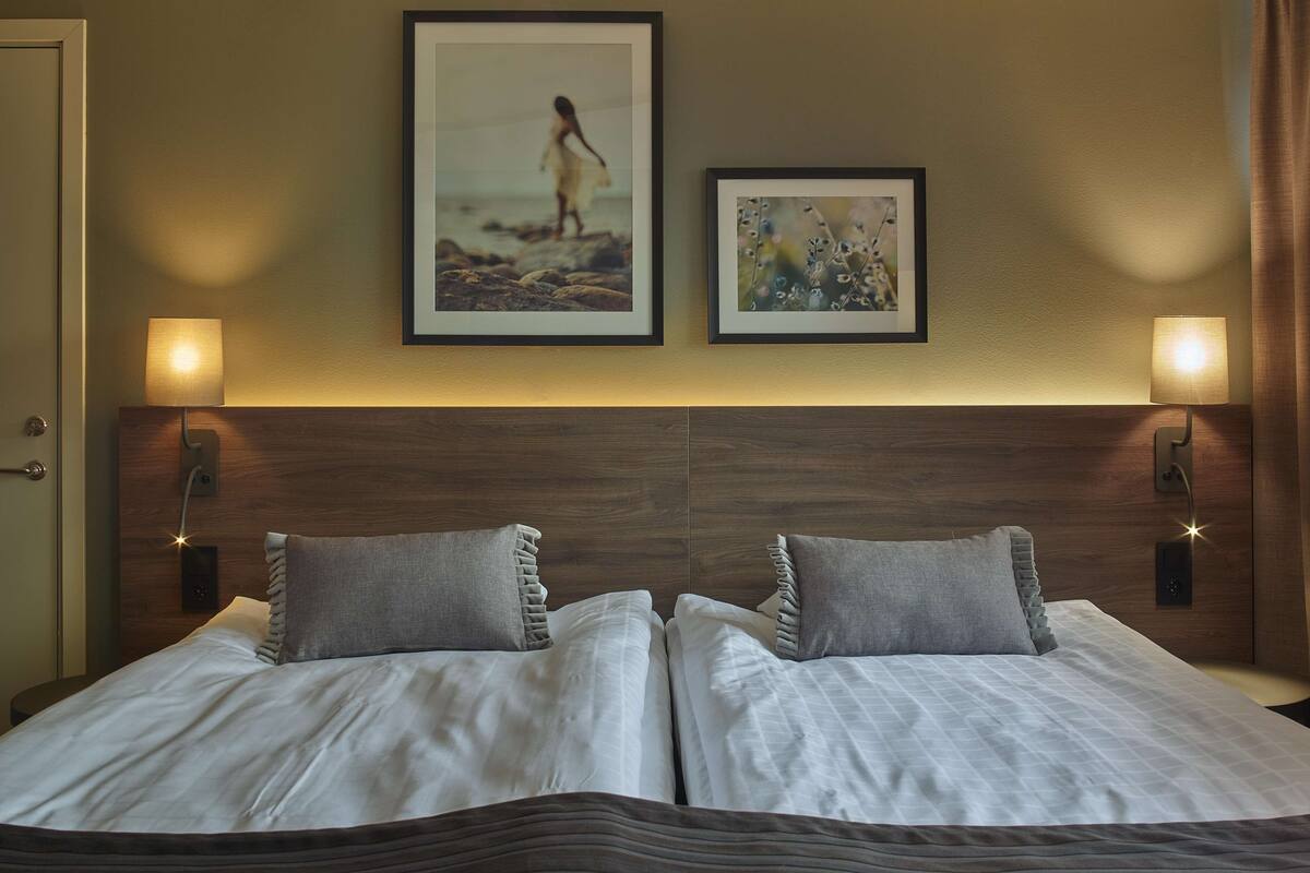 Una cama doble con dos almohadas, un cabecero de madera, dos lámparas fijadas a la pared y dos cuadros enmarcados.