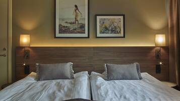 A double bed with two pillows, a wooden headboard, two wall-mounted lamps, and two framed pictures on the wall.