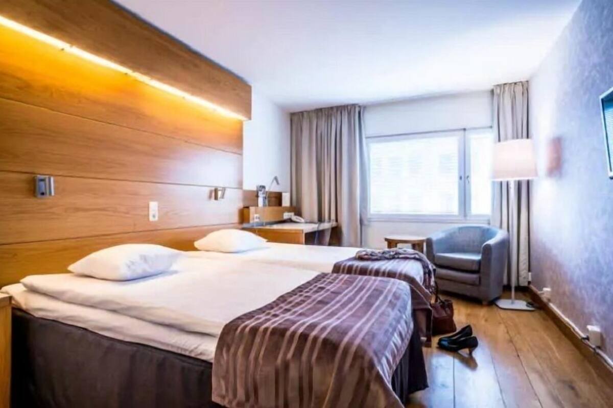 Habitación de hotel con dos camas, un escritorio, una silla y un ventanal con cortinas.