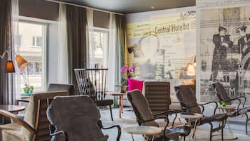 A modern hotel lobby with comfortable seating, large windows, and a wall decorated with historical posters.