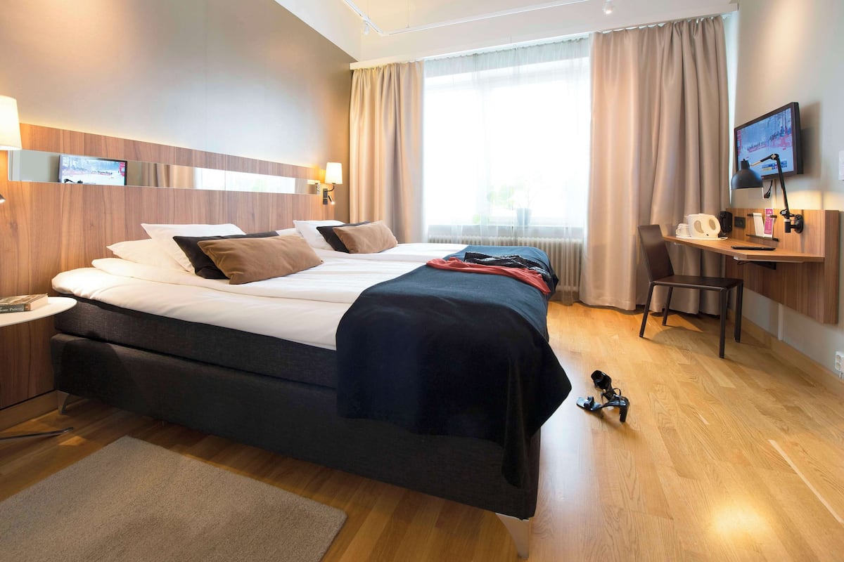 Una habitación de hotel con una cama grande, un escritorio con silla, televisión y ventana con cortinas.