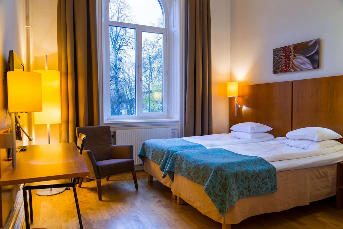 Habitación de hotel con cama, escritorio, silla, ventana con cortinas y un cuadro en la pared.