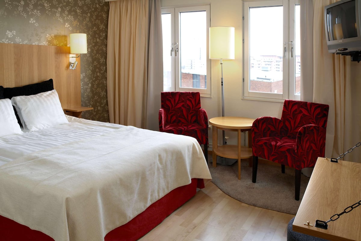 Habitación de hotel con cama, una mesa, dos sillas, televisión y ventana con cortinas.