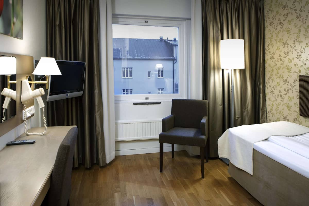 Una habitación de hotel con cama, una silla, un escritorio con televisor y una ventana que da a edificios.