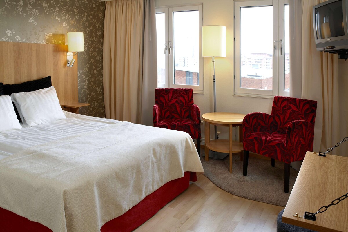 Ett hotellrum med en säng, ett skrivbord, två stolar, en tv och ett fönster.