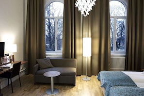 Habitación de hotel con sofá, una mesita redonda, cama con ropa de cama estampada, lámpara de pie y ventanas grandes con cortinas.