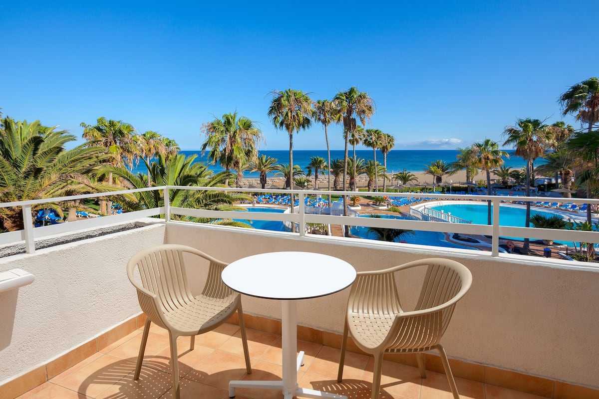 A balcony with two chairs and a small table, overlooking a pool and beach area with palm trees and a clear blue sky.
