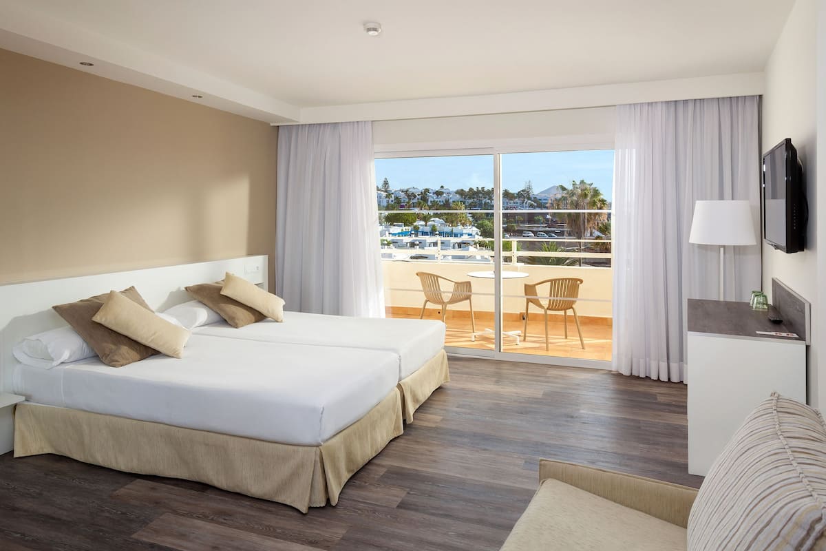 A hotel room with a large bed, a TV, a desk, and a balcony with a view of the city.