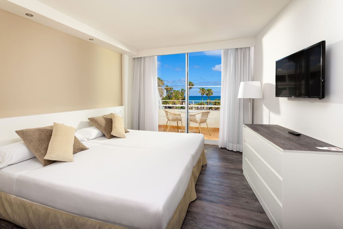 A hotel room with a bed, a TV, and a balcony with a view of the ocean.