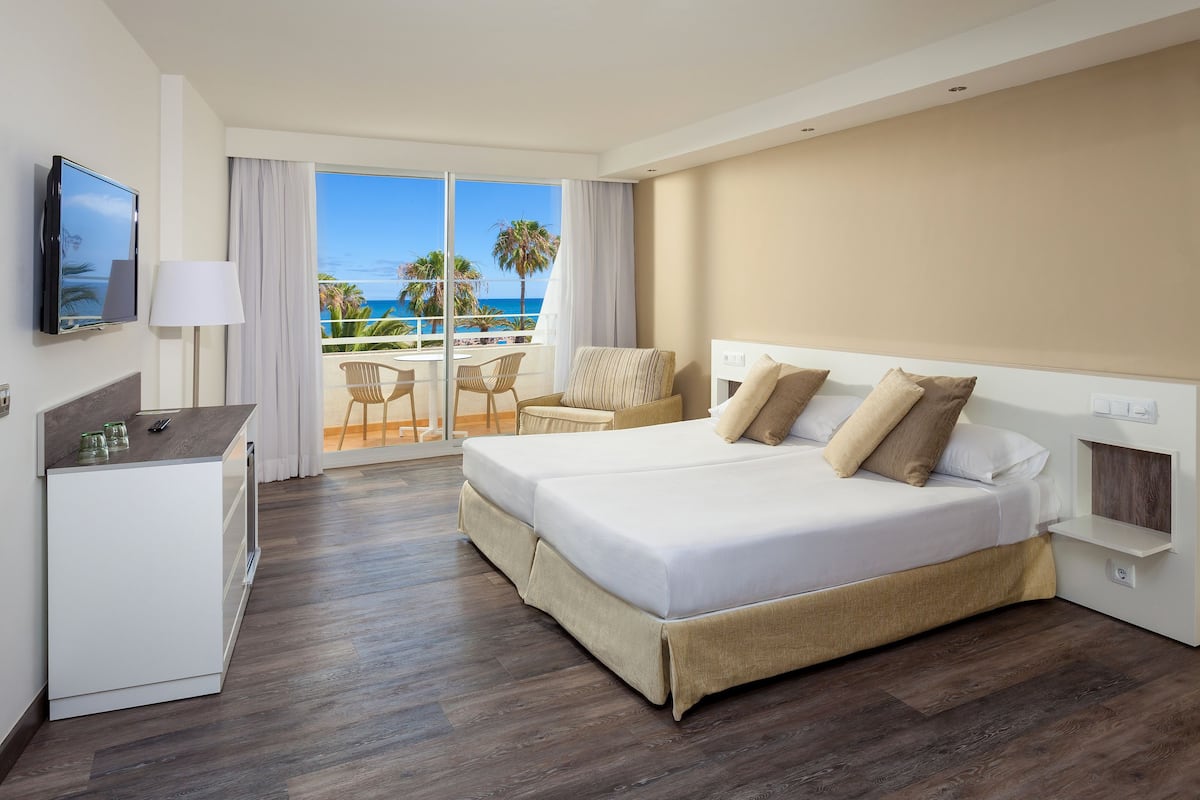 A hotel room with a large bed, a desk with a TV, a balcony with a view of the ocean, and a chair.