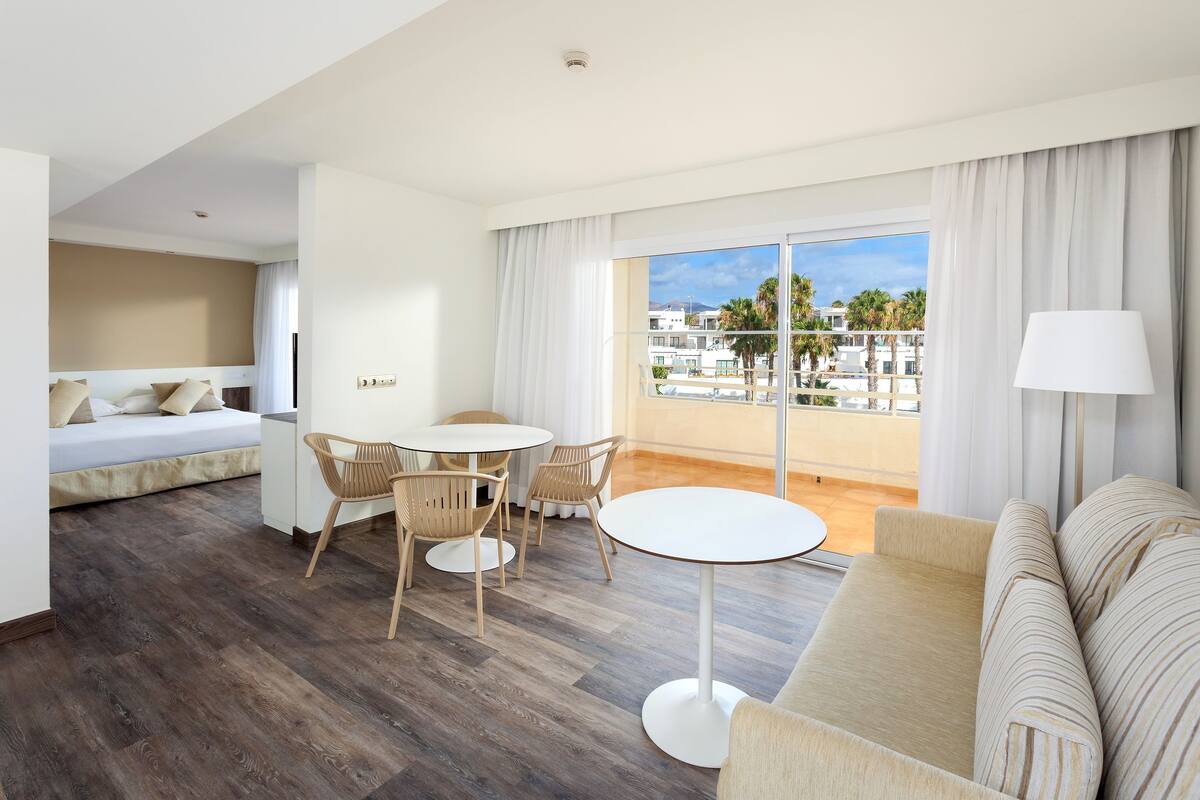 A modern hotel room with a balcony, a sofa, a dining table with chairs, and a bed.