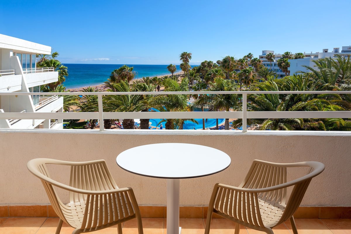 A balcony with two wicker chairs and a small round table, overlooking a beach with palm trees and a clear blue sky.
