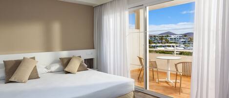 A hotel room with a large bed, a balcony with a table and chairs, and a view of buildings and mountains.