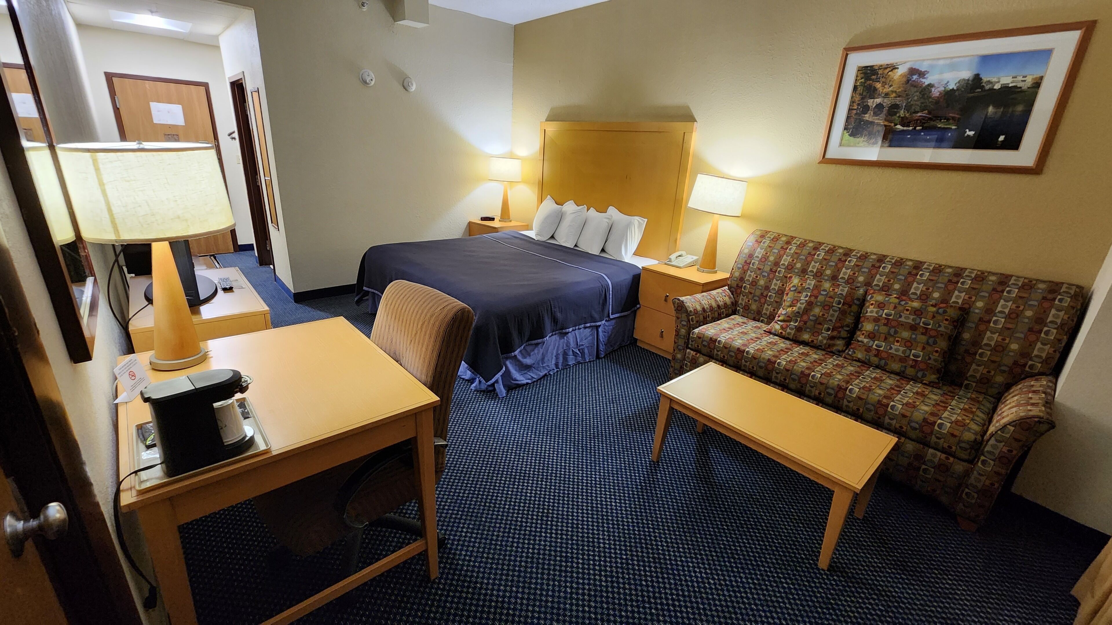 Ein Hotelzimmer mit einem Bett, einer Couch, einem Schreibtisch und einem Stuhl. Zwei Nachttische mit Lampen und ein gerahmtes Bild an der Wand.