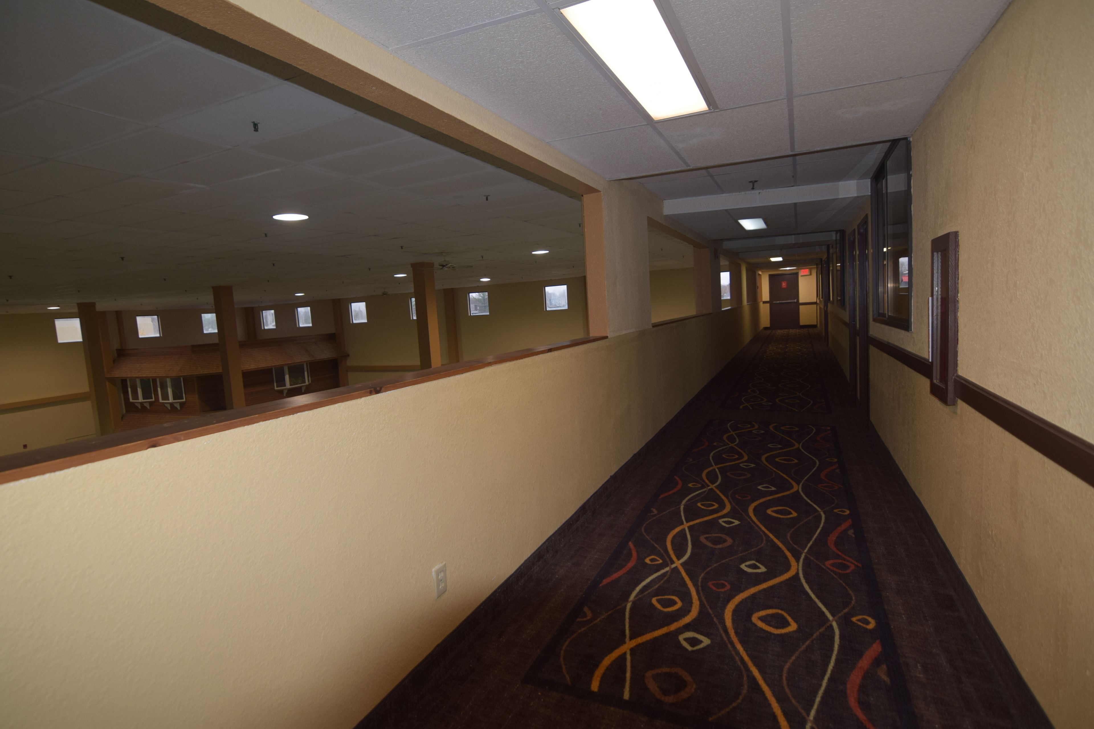 A long, narrow hallway with a patterned carpet, beige walls, and a ceiling with fluorescent lights.