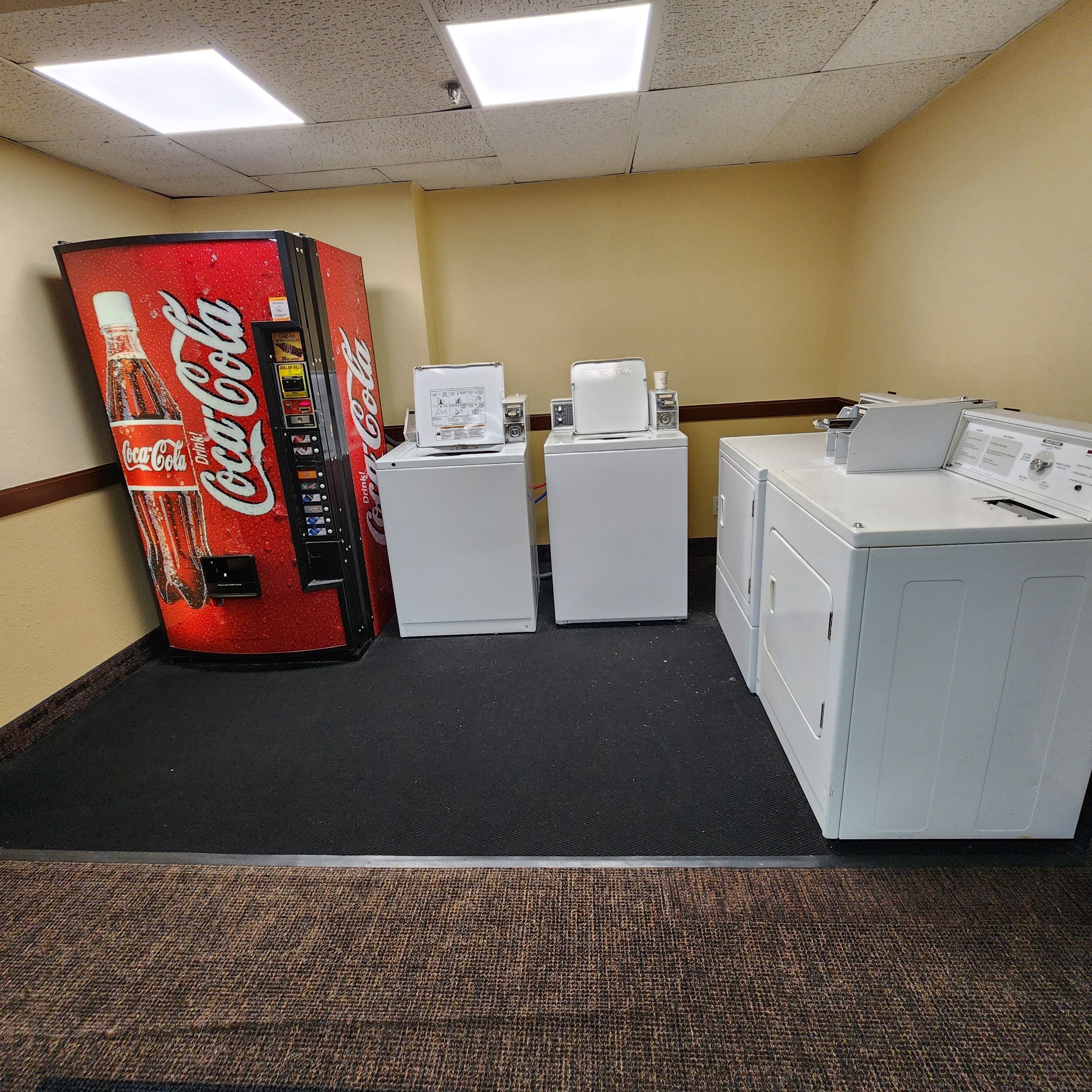 A room with a Coca-Cola vending machine, two dryers, and a washing machine.