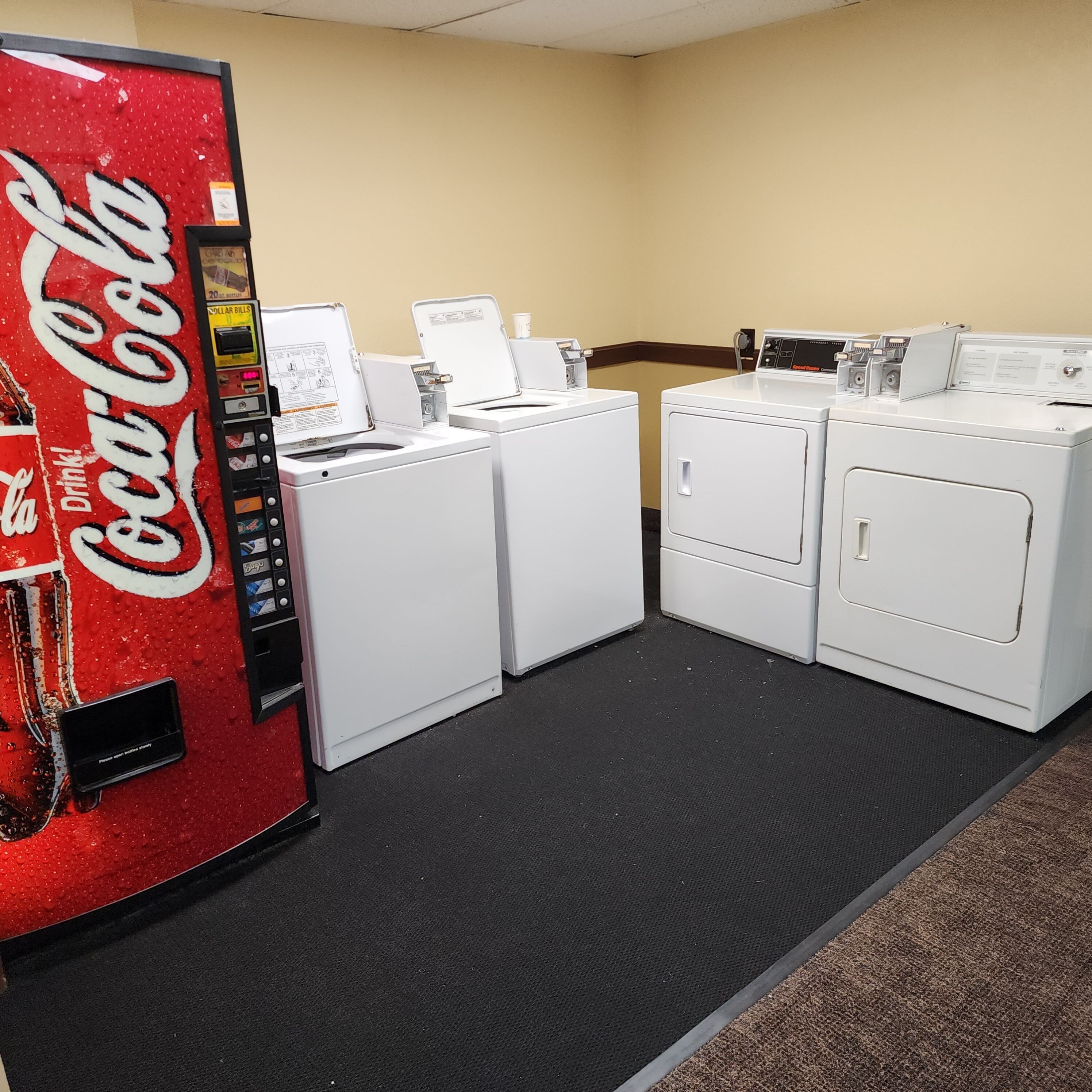 A laundromat with a Coca-Cola vending machine, multiple washers and dryers, and a carpeted floor.