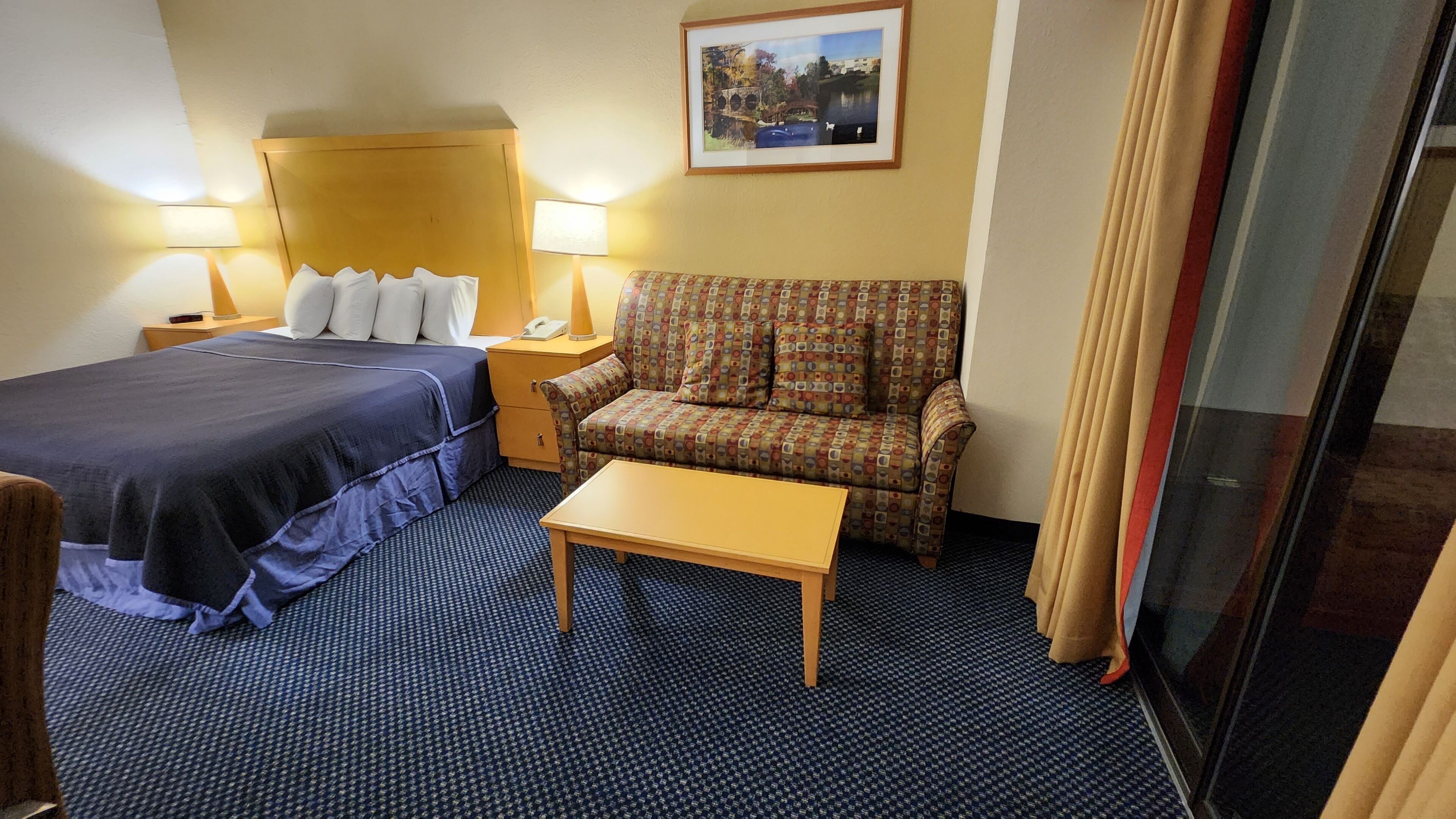 A hotel room with a bed, a sofa, a small table, and a nightstand with a lamp.