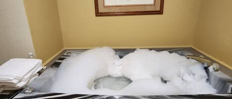A bathtub filled with bubbles, a framed picture on the wall, and a stack of white towels on the side.