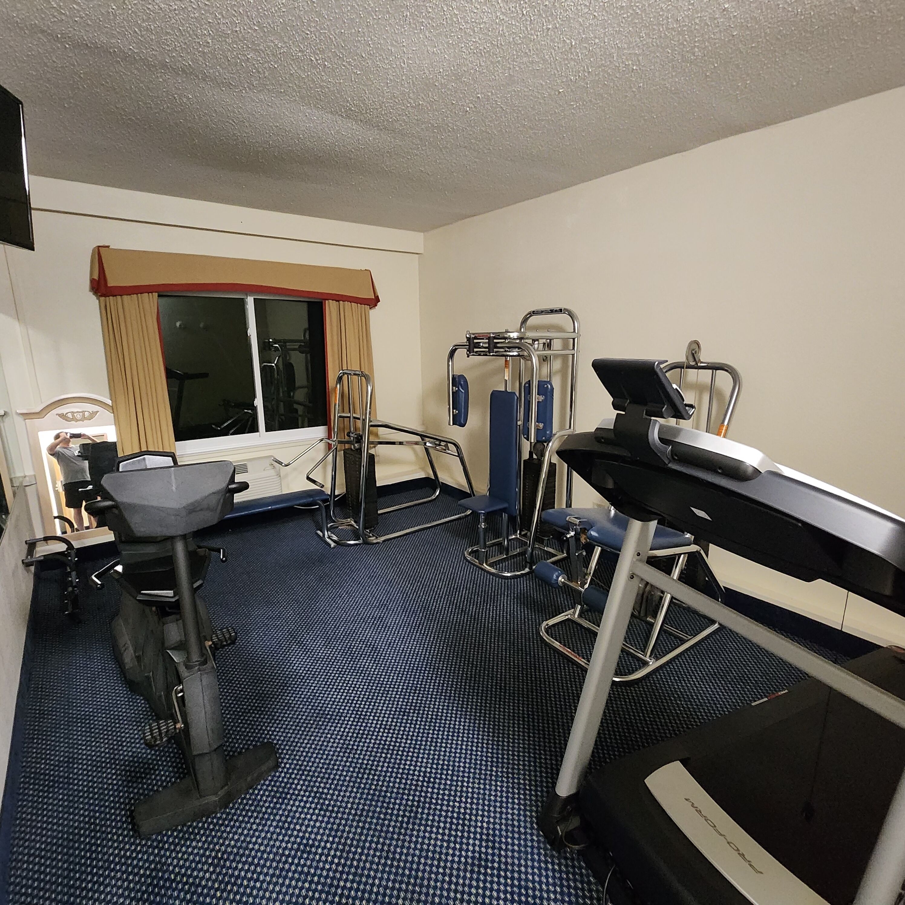 A hotel room with a treadmill, exercise equipment, and a window with curtains.