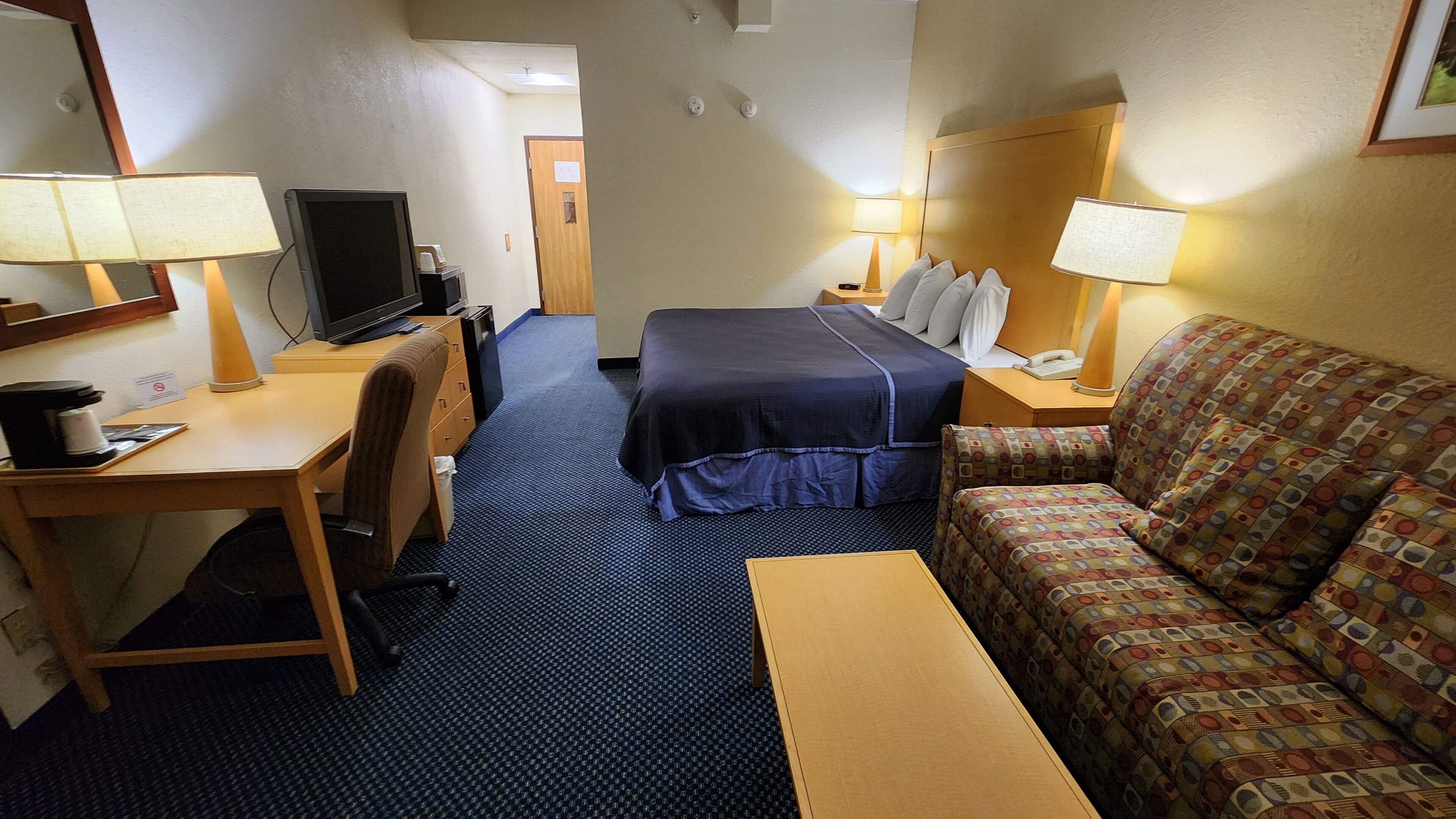 Hotellihuone, jossa on sänky, työpöytä tietokoneella, tuoli, sohva ja kaksi yöpöytävalaisinta.