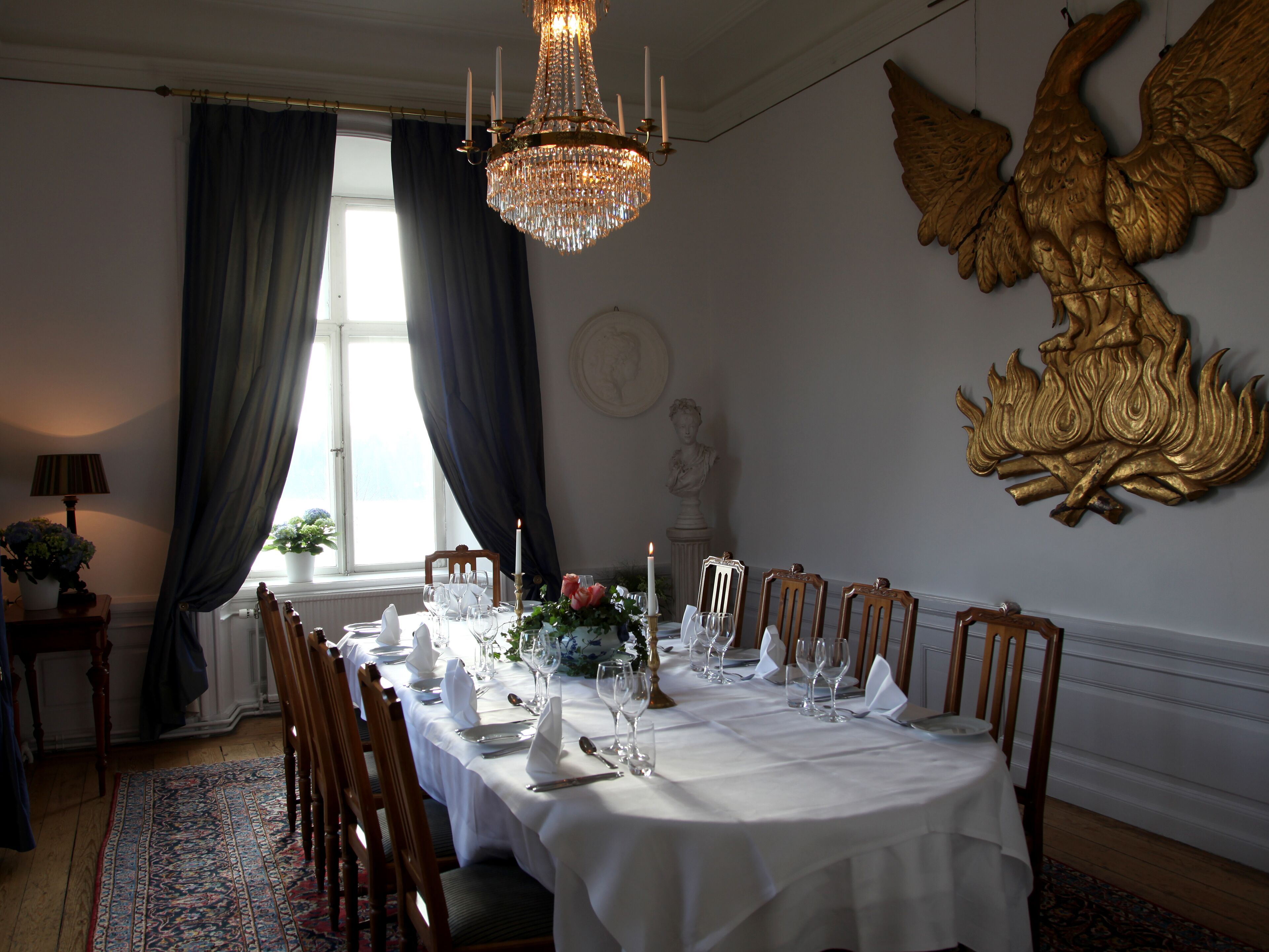 A formal dining room with a long table set for a meal, a chandelier, and a large golden winged sculpture on the wall.