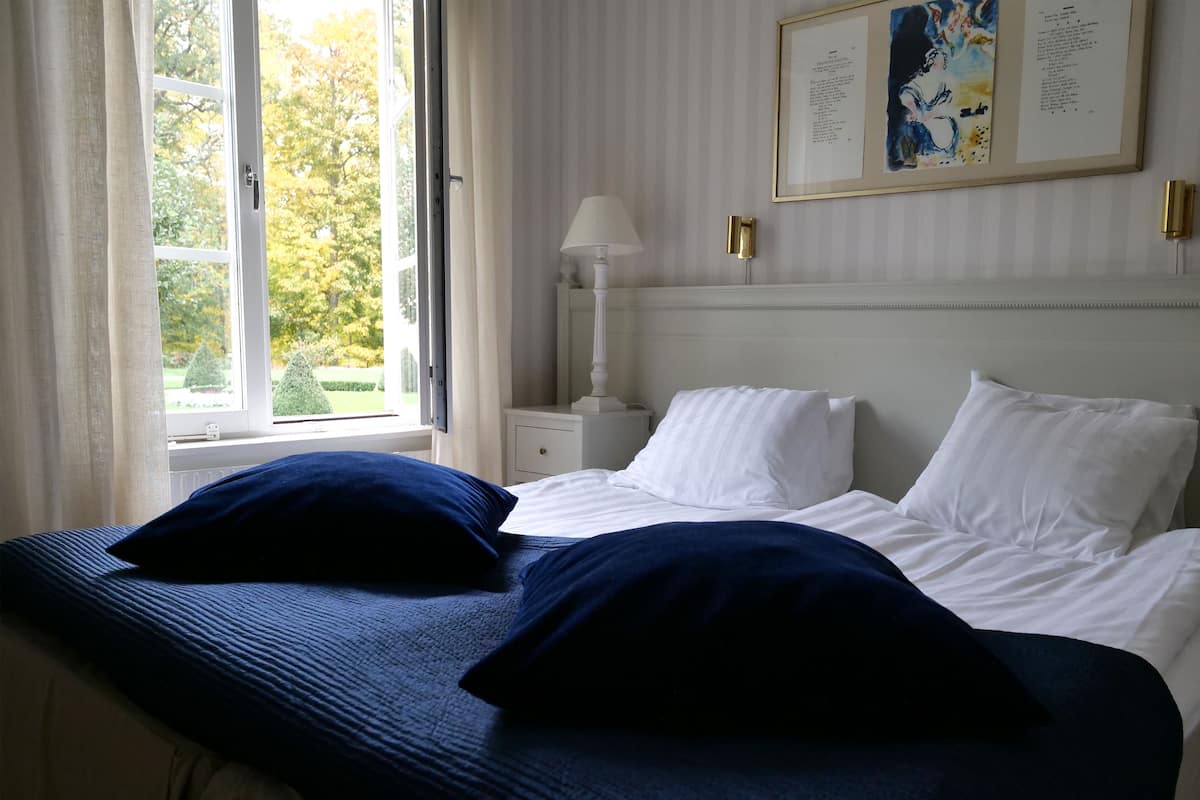 A hotel room with a large bed, white bedding, a nightstand with a lamp, and a window with curtains.