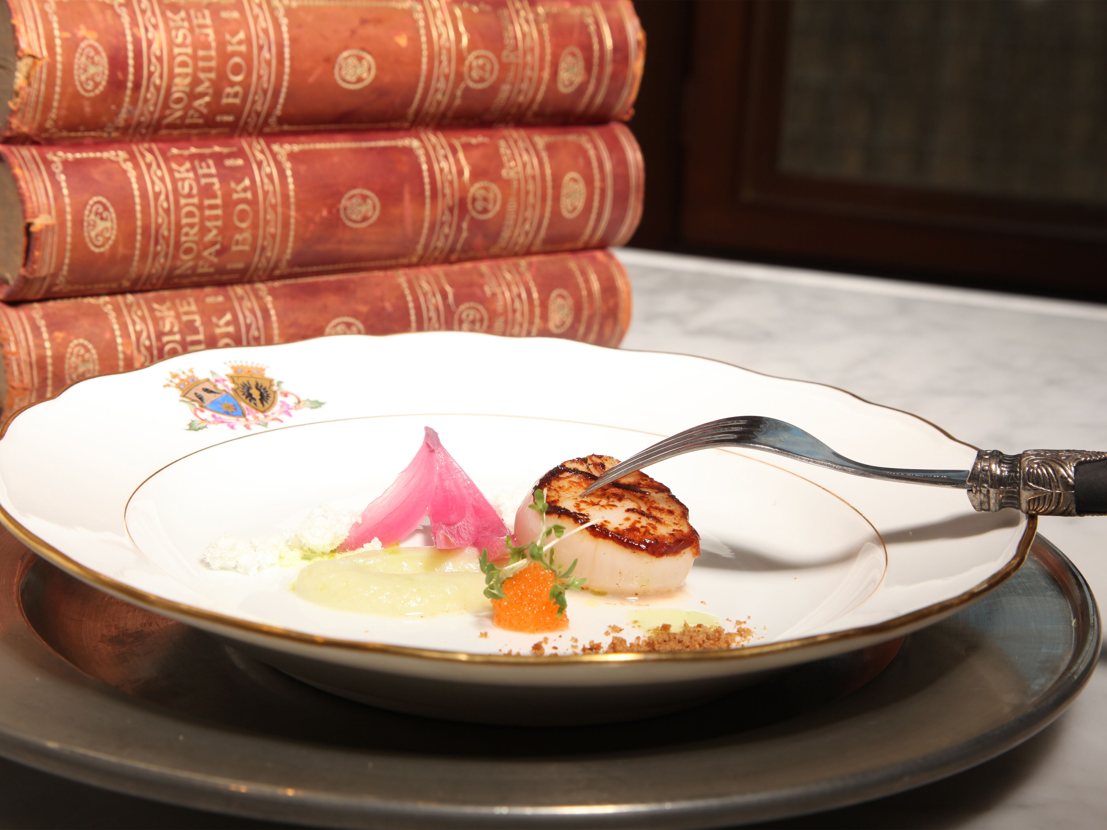 A plate with a seared scallop, a pink vegetable, and a garnish, accompanied by a fork on a white plate with gold trim.