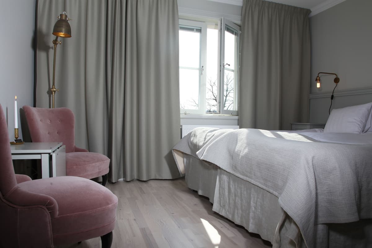 A bedroom with a bed, a chair, a nightstand, a lamp, and a window with curtains.