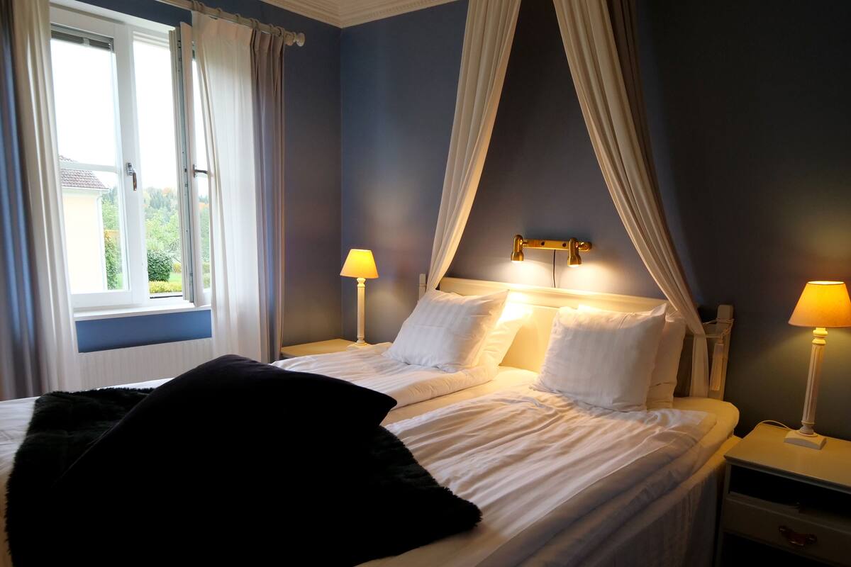 A bedroom with a bed, two bedside tables, a window with curtains, and a canopy bed.