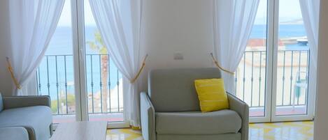 A living room with a grey sofa, a yellow pillow, and a wooden coffee table. The room has a yellow and white chevron-patterned floor and large windows with white curtains.