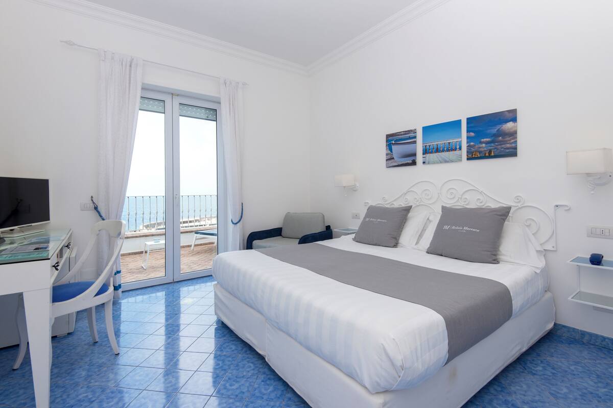 A hotel room with a bed, a desk, a chair, a balcony with a view, and a wall with three pictures.