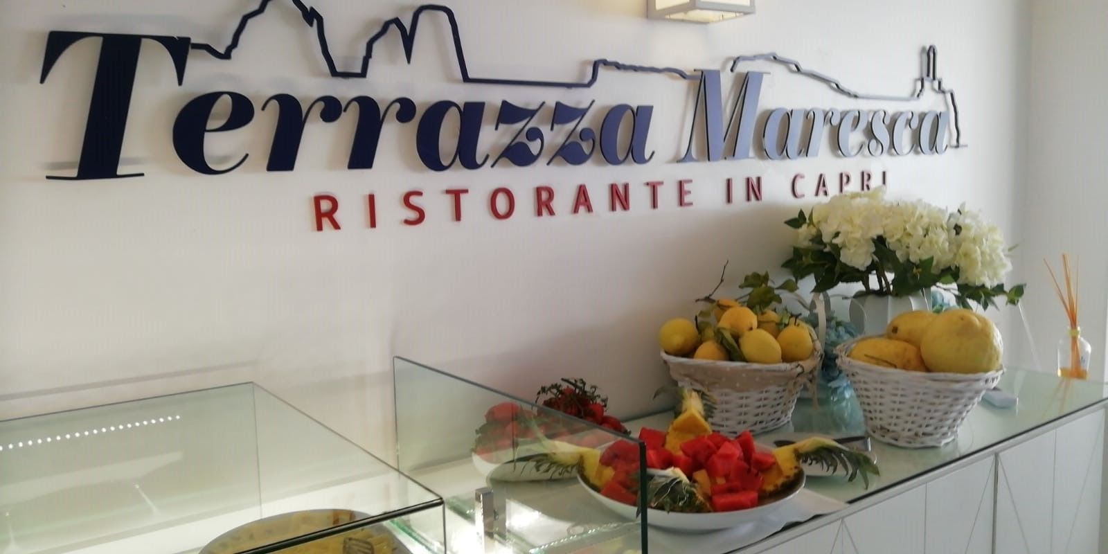 Une enseigne de restaurant portant le nom « Terrazza Marcesca » et la formule « Ristorante in Capri ». On peut voir des paniers de fruits et un vase rempli de fleurs sur le comptoir.