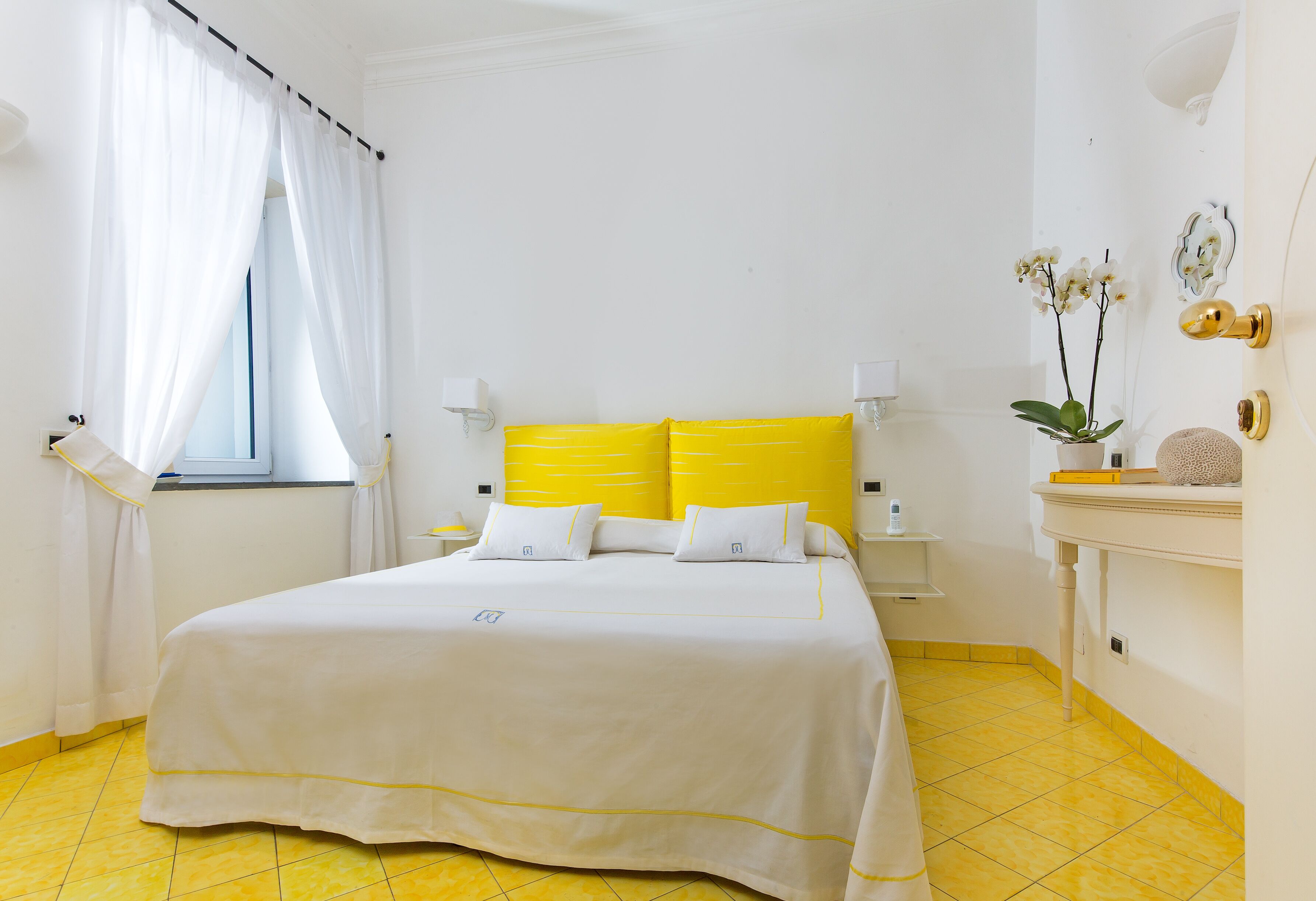Habitación de hotel con cama, cortinas blancas, mesita de noche, lámpara, un florero con flores y suelo de baldosas amarillas.