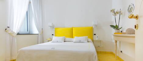 Habitación de hotel con cama, cortinas blancas, mesita de noche, lámpara, un florero con flores y suelo de baldosas amarillas.
