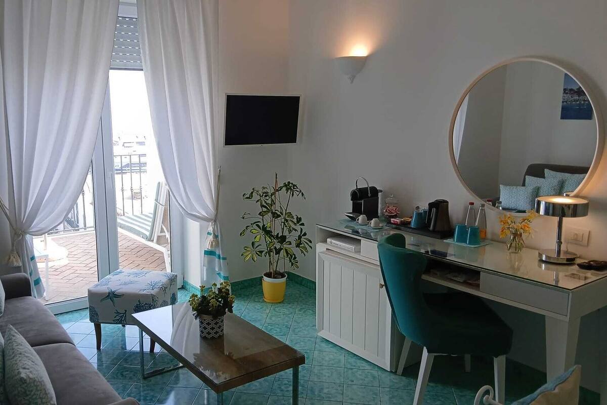 A compact living space with a sofa, a small table, a desk with a chair, a potted plant, a flat-screen TV, and a balcony with a view.