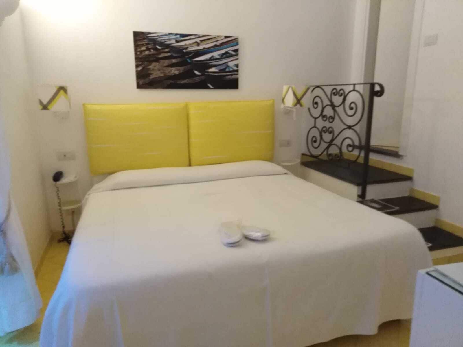 A bedroom with a bed, a yellow headboard, a painting on the wall, a staircase with a black railing, and a white bedside table.