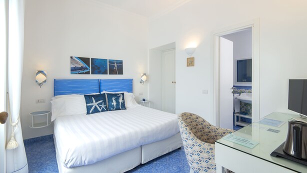 A hotel room with a bed, a chair, a desk with a kettle, and artwork on the wall.