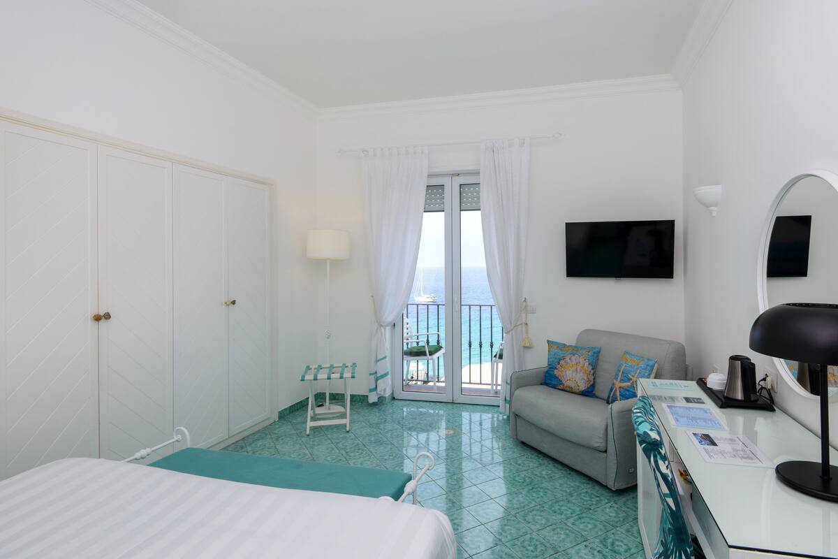 A hotel room with a bed, a desk with a lamp, a sofa with cushions, a TV on the wall, and a balcony with a view of the sea.