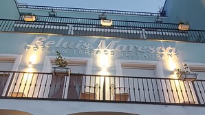 A hotel entrance with a sign reading "Jelais Marecasa Hotel & Restaurant," featuring a balcony with potted plants and a warm, inviting interior.