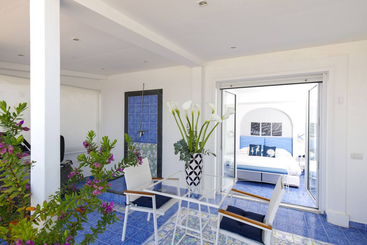 A modern hotel room with a glass table, white chairs, a bed with blue bedding, and a view through a glass door.