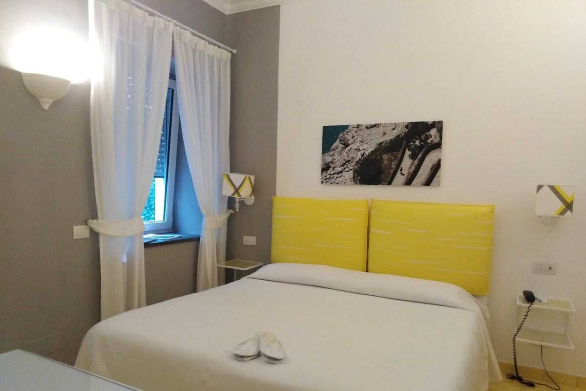 A bedroom with a bed, a window with curtains, a bedside table, a wall-mounted phone, and a picture on the wall.
