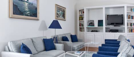 Ein modernes Wohnzimmer mit einem grauen Sofa, blauen Sessel und einem Couchtisch, sowie einem Bücherregal mit einem Fernseher.